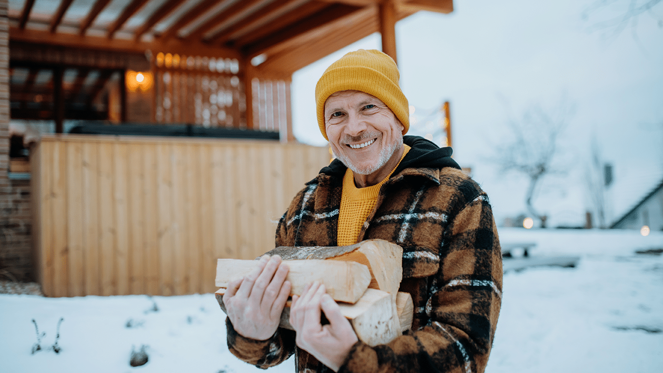 Henkilö pitelee ulkona halkoja sylissään, katsoo suoraan kemraan ja hymyilee leveästi
