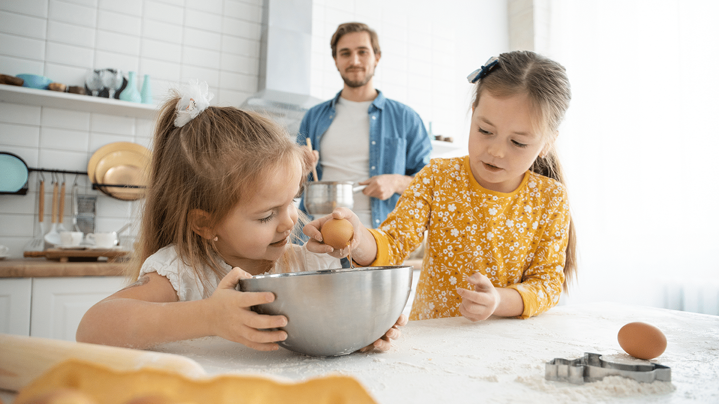 Kaksi lasta leipoo. Vanhempi seisoo taustalla.