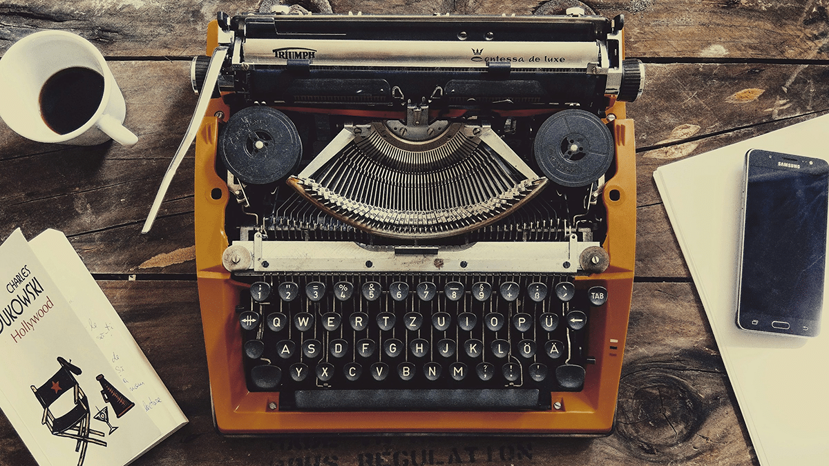 Vanha kirjoituskone, ympärillä kahvikuppi, kirja ja kännykkä.