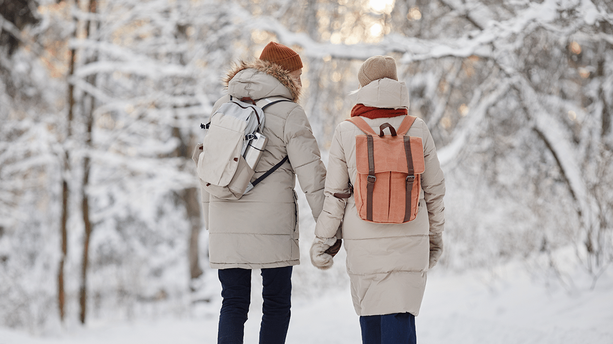 Takaapäin otettu kuva, jossa kaksi ihmistä kävelee talvisessa metsässä käsi kädessä.